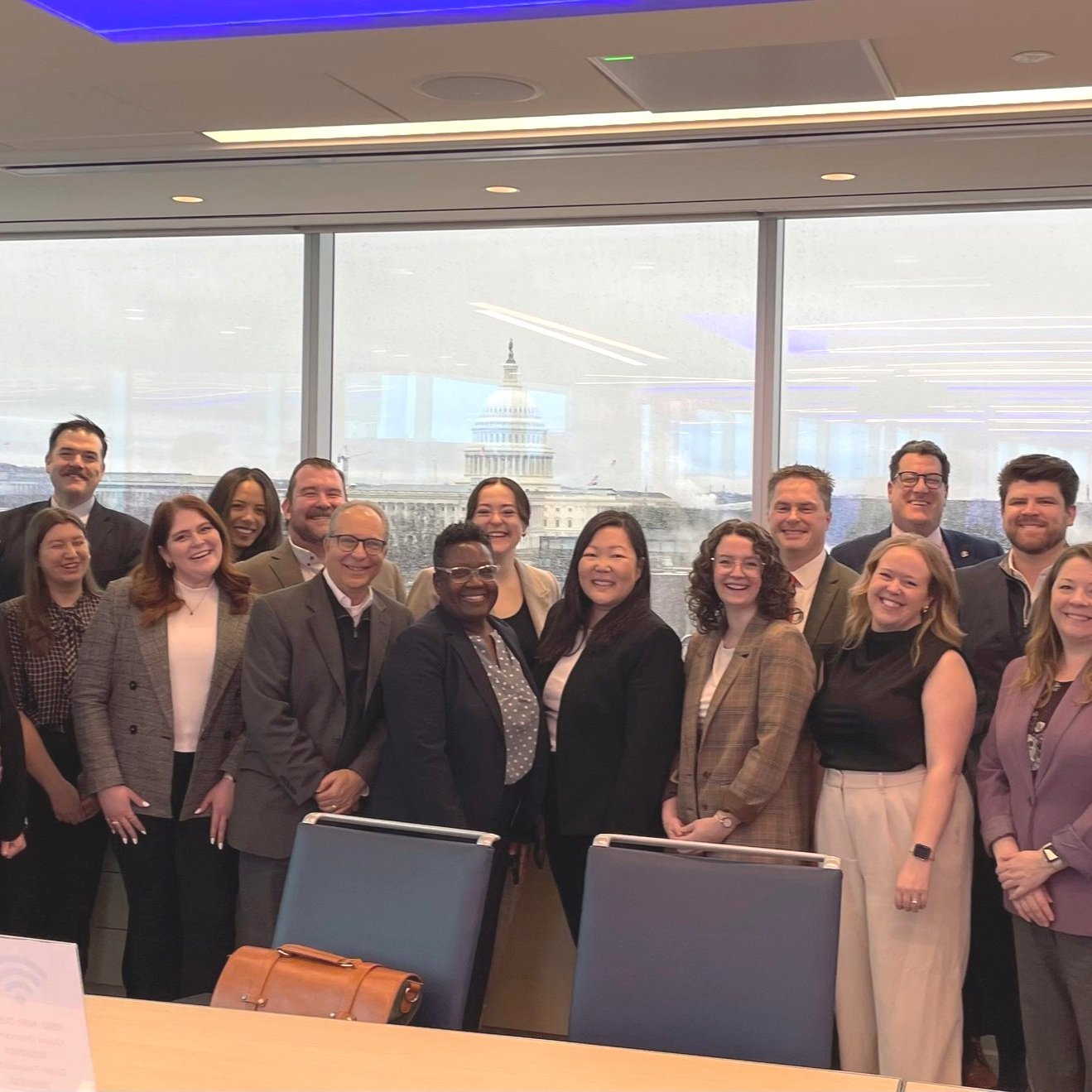 Group of people in business attire smiling in an office with the U.S. Capitol building visible through the window.