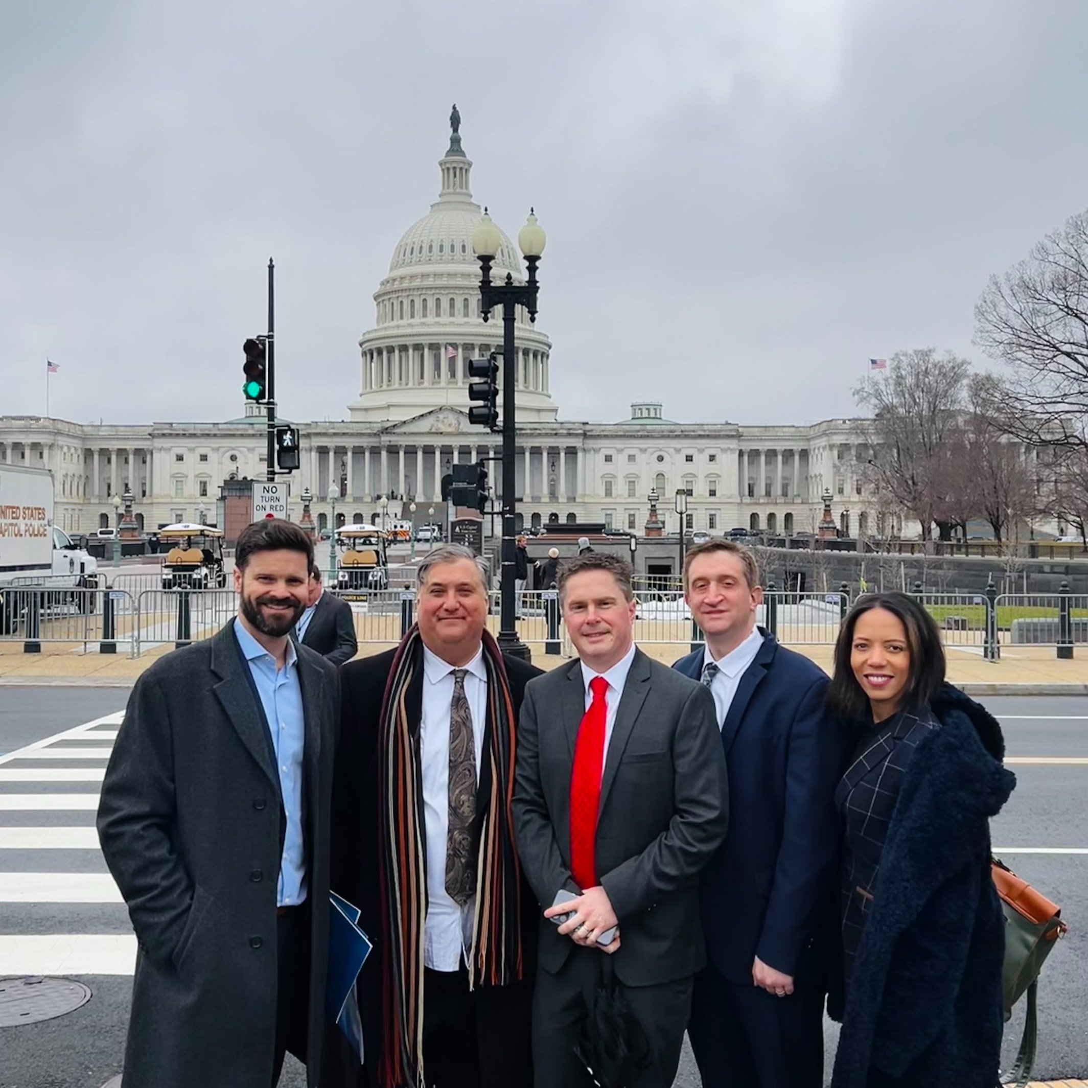 Group of five people posing in front of the U.S. Capitol building on a cloudy day.